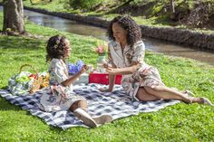 two women sitting on a blanket in the grass near a river and picnicking together