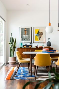 a dining room table with yellow chairs and art on the wall above it, along with potted plants