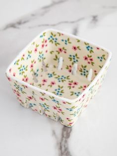 a small square candle holder with flowers on it sitting on a marble countertop next to candles