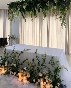 an arrangement of flowers and candles on a white table cloth with greenery hanging from the ceiling