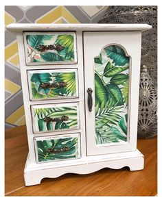 a white cabinet with tropical leaves painted on the doors and drawers is sitting on a wooden table