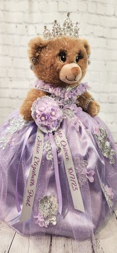 a brown teddy bear wearing a purple dress and tiara sitting on top of a wooden table