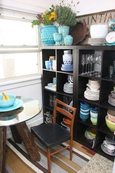 a dining room table with plates and bowls on it next to a shelf filled with dishes