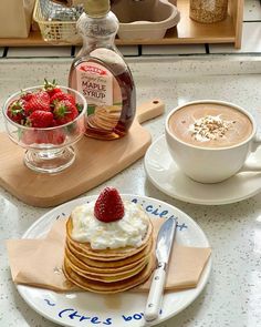 pancakes with whipped cream and strawberries are on a plate next to a cup of cappuccino