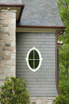 a round window on the side of a gray brick house with green trees in the background