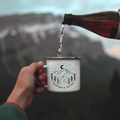 a person is pouring water into a coffee mug with mountains in the background and trees on it