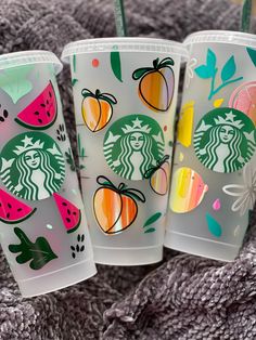 three starbucks cups with fruit designs on them are sitting on a gray blanket and one has a straw in it