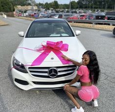 a woman kneeling next to a white car with a pink bow on it's hood