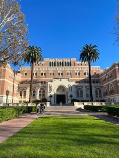 two people standing in front of a large building with palm trees on the side walk