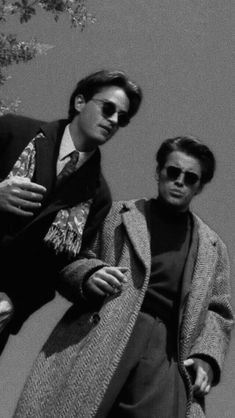 two men standing next to each other in front of a tree wearing sunglasses and coats