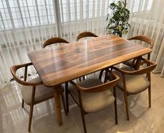 a wooden table with chairs around it in front of sheered curtains and a potted plant