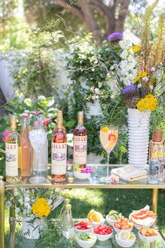 an assortment of alcohol bottles and glasses on a table with flowers in the back ground