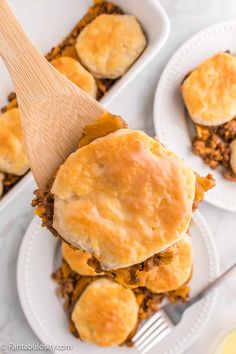 two white plates topped with biscuits covered in meat and cheese next to a wooden spatula