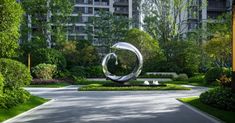 a large metal sculpture sitting in the middle of a lush green park next to tall buildings