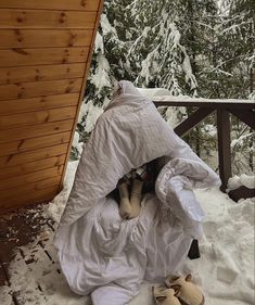 a dog wrapped in a blanket on top of snow covered ground next to a wooden cabin