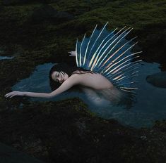 a woman is floating in the water with wings on her head and body, surrounded by moss