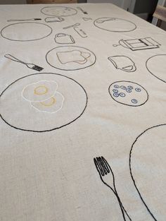 an embroidered tablecloth with various food items and utensils on it, along with a wooden chair