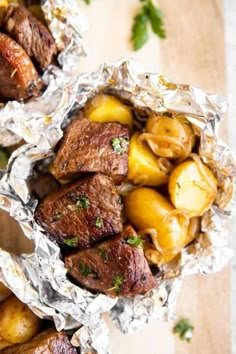 two foil packets filled with meat and potatoes on top of a wooden cutting board next to parsley