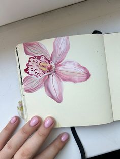 a woman's hand with pink nail polish holding an open book that has a flower painted on it