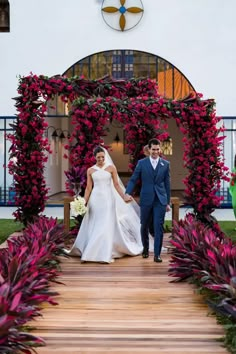 a bride and groom walking down a wooden walkway in front of a building with purple flowers