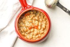 a red bowl filled with beans next to a spoon and pepper shaker on a white surface