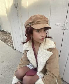 a woman sitting on the ground wearing a coat and hat with her legs crossed, in front of a garage door