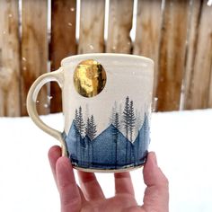 a hand holding a coffee cup with trees on it in the snow near a fence