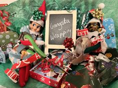 an assortment of christmas presents and elfs with a chalkboard on the wall behind them