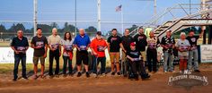 a group of people standing next to each other in front of a metal fence and baseball field