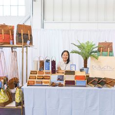 a woman sitting behind a table with bags and purses on it, smiling at the camera