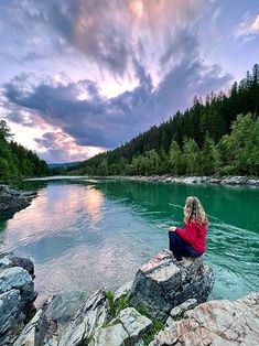 a woman sitting on top of a large rock next to a river under a cloudy sky