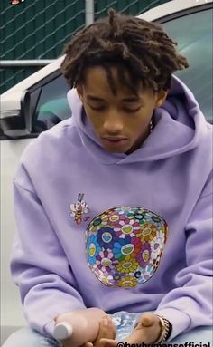 a young man sitting on top of a white car wearing a purple hoodie and looking at his cell phone