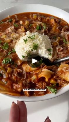 a person holding a spoon in a bowl filled with chili and cheese covered meat stew