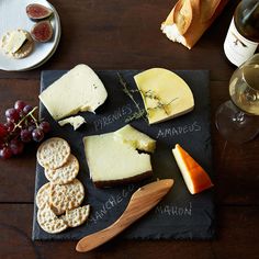 various cheeses and crackers are on a slate board with wine, bread, and grapes