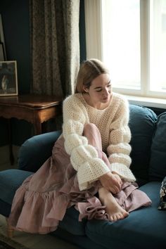 a woman sitting on top of a blue couch in front of a window wearing a sweater