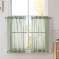 a window with green sheer curtains in front of a white wall and wooden dresser next to it