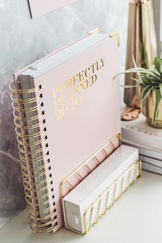 a pink notebook with gold foil lettering on it next to other office supplies and plants