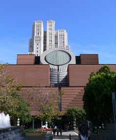 a large clock on the side of a building in front of trees and people walking