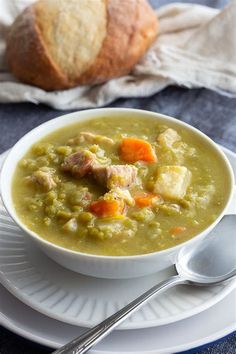 a bowl of soup with meat and vegetables on a white plate next to a loaf of bread