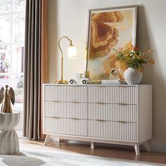 a white dresser sitting next to a window in a room with a painting on the wall