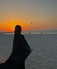 a woman sitting on top of a wooden bench near the ocean at sunset with kites flying in the sky