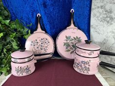 three pink ceramic pots and pans with flowers painted on them sitting next to a blue curtain