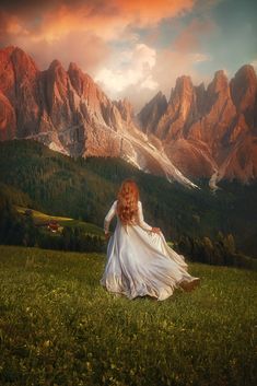 a woman in a white dress sitting on top of a lush green field next to mountains