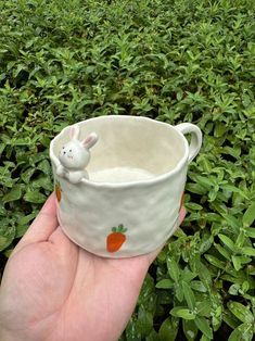 a hand holding a white cup with a bunny in it on top of green bushes