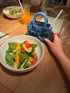 a salad in a white bowl on a table next to a hand holding a blue bag