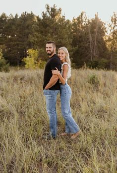 a man and woman standing in tall grass with their arms around each other looking into the distance