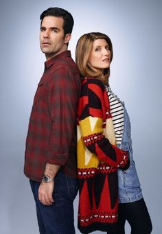 a man and woman standing next to each other in front of a gray background wearing sweaters