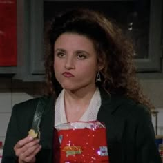 a woman holding a bag of chips in her hand and looking at the camera while standing in a kitchen