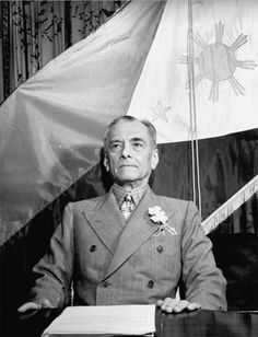 an older man sitting at a desk in front of two flags