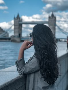 a woman with long black hair standing next to the water and looking at the tower bridge